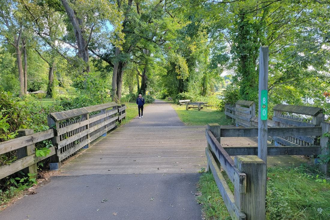 A wooden boardwalk on the Farmington River Trail at Collinsville, Connecticut, with a picnic table on the grass and the river visible through the trees.