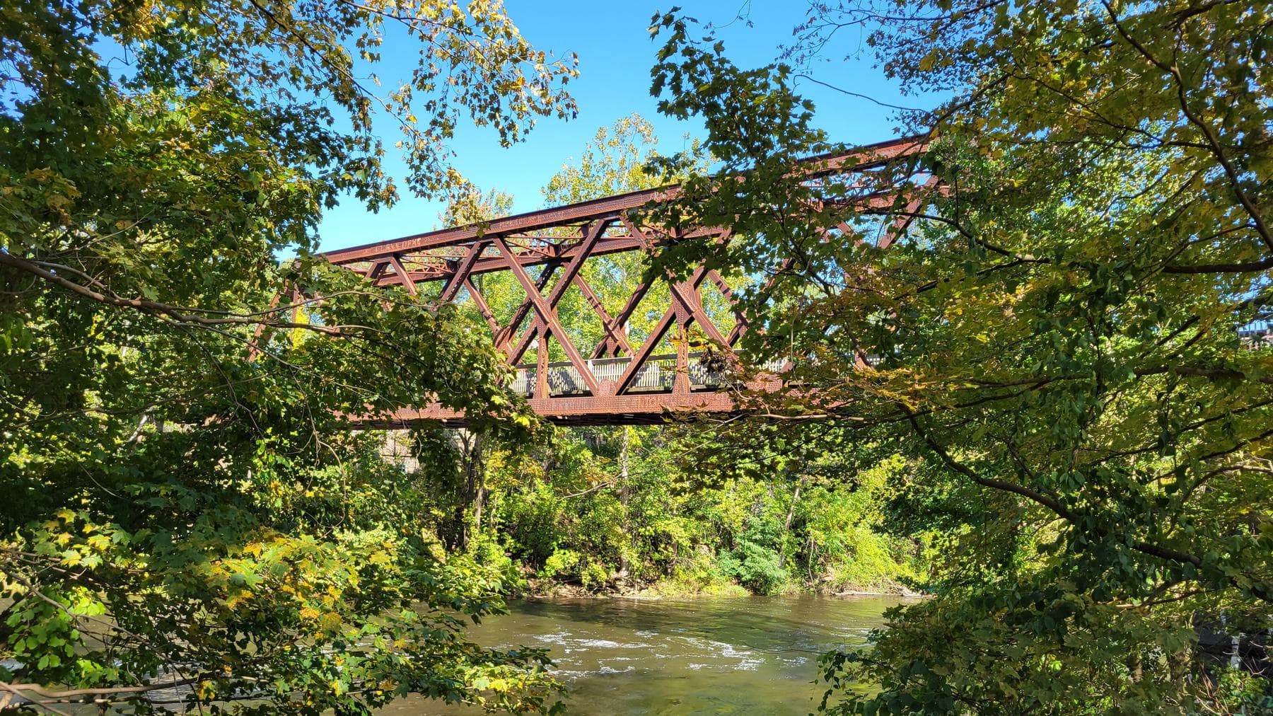 An iron truss rail bridge carrying the Farmington River Trail over the Farmington River at Collinsville, Connecticut, framed by green and gold leaves under a blue sky.